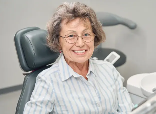 Smiling mature woman in dental chair wearing glasses and blue striped shirt.