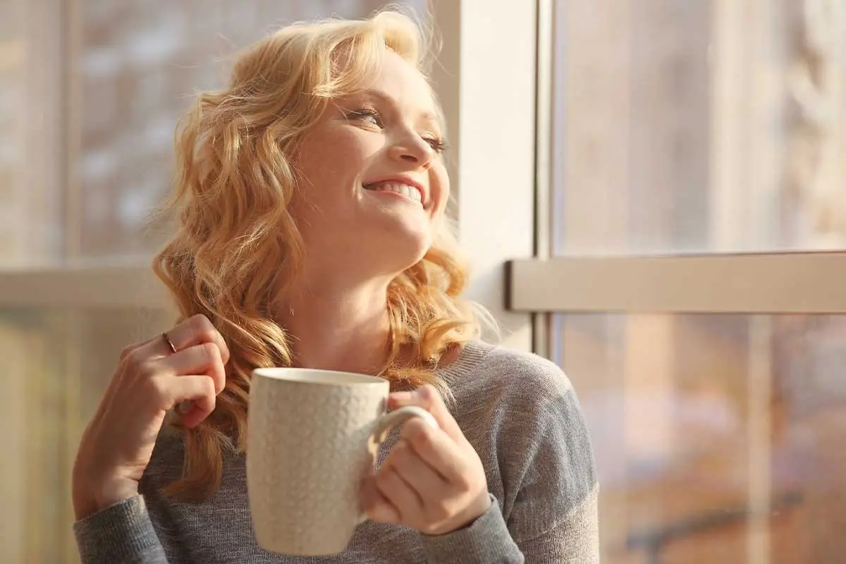 Woman with red hair smiling while holding a warm beverage by a window.