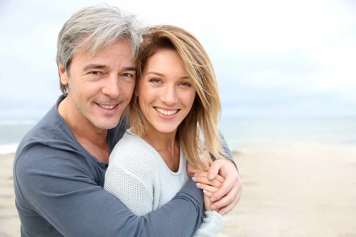 Happy middle-aged couple smiling on beach, representing healthy lifestyle and dental wellness.