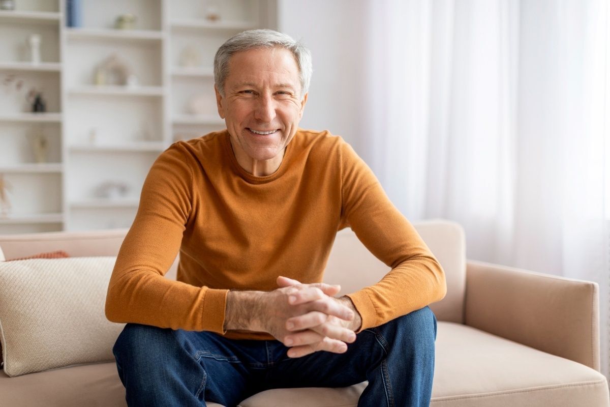 Smiling senior man in orange sweater sitting on couch in modern dental office.