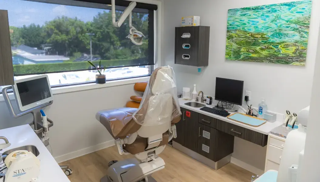 Modern dental treatment room with patient chair, overhead light, digital monitor, sink station, and large window overlooking.