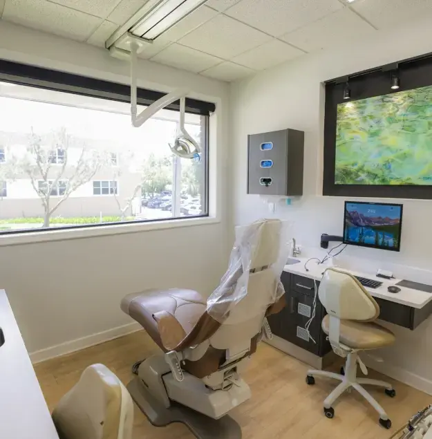 Modern dental treatment room with comfortable patient chair, overhead lighting, multiple monitors, and large window with nat.